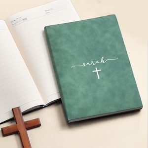 Op de afbeelding: Een saliegroen notitieboek met een getextureerde kaft waarop de naam Sarah en een kruissymbool in het wit zijn gedrukt. Het notitieboek ligt naast een open gelinieerde dagboekpagina en een klein, donkerbruin houten kruis op de ondergrond.