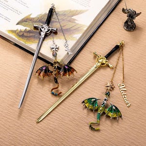May include: Sword-shaped metal bookmarks with dragon charms. One bookmark is silver with a black handle and a dragon with orange, purple, and black wings. The other is gold with a black handle and a green and yellow dragon.