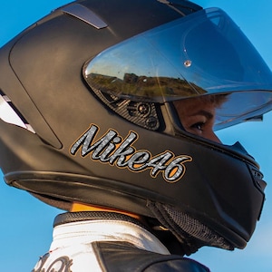 以下が含まれることがあります： 透明なバイザーが付いた黒いオートバイのヘルメット。ヘルメットには「Mike46」の文字が銀色とオレンジ色で書かれています。ヘルメットをかぶっている人は、白と黒のジャケットを着ています。