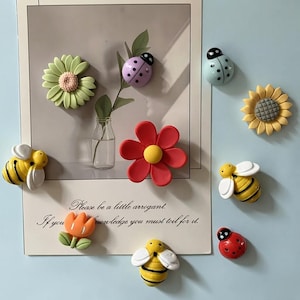 Könnte beinhalten: Eine Sammlung dekorativer Magnete mit drei gelb-schwarzen Bienen, zwei Marienkäfern in Hellblau und Rot, einem hellgrünen Gänseblümchen, einer gelben Sonnenblume, einer roten Blume und einer orangefarbenen Tulpe, angeordnet um eine Karte mit Text.