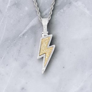 Puede incluir: Collar de cadena plateada con un colgante en forma de rayo. El rayo tiene un centro dorado y está bordeado con pequeñas piedras preciosas transparentes. La cadena tiene un diseño de cuerda trenzada.