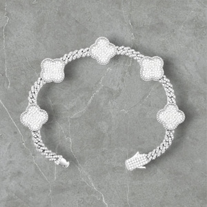 Op de afbeelding: Een zilveren armband met een klaverontwerp. De armband heeft een schakelband met vijf klavervormige bedels, elk bezet met kleine, sprankelende stenen. De sluiting is open en toont de ingewikkelde details van het ontwerp.