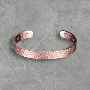 Op de afbeelding: Een koperkleurige armband met een gehamerde textuur. Het open manchetontwerp heeft afgeronde uiteinden met kleine cirkelvormige inkepingen. De armband heeft een warme, metallic tint en het oppervlak heeft een putjespatroon.