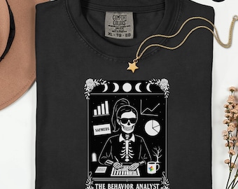 May include: Black Comfort Colors t-shirt with a graphic of a skeleton in a business setting, with the text "THE BEHAVIOR ANALYST". A gold star necklace and a hat are also visible. The shirt has a tag that says XL-TG-EG.