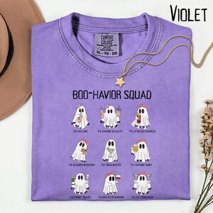 May include: A violet Comfort Colors t-shirt with the text "BOO-HAVIOR SQUAD" and illustrations of ghosts. Each ghost is depicted with a different accessory and title. A gold necklace with a star pendant is draped over the shirt.