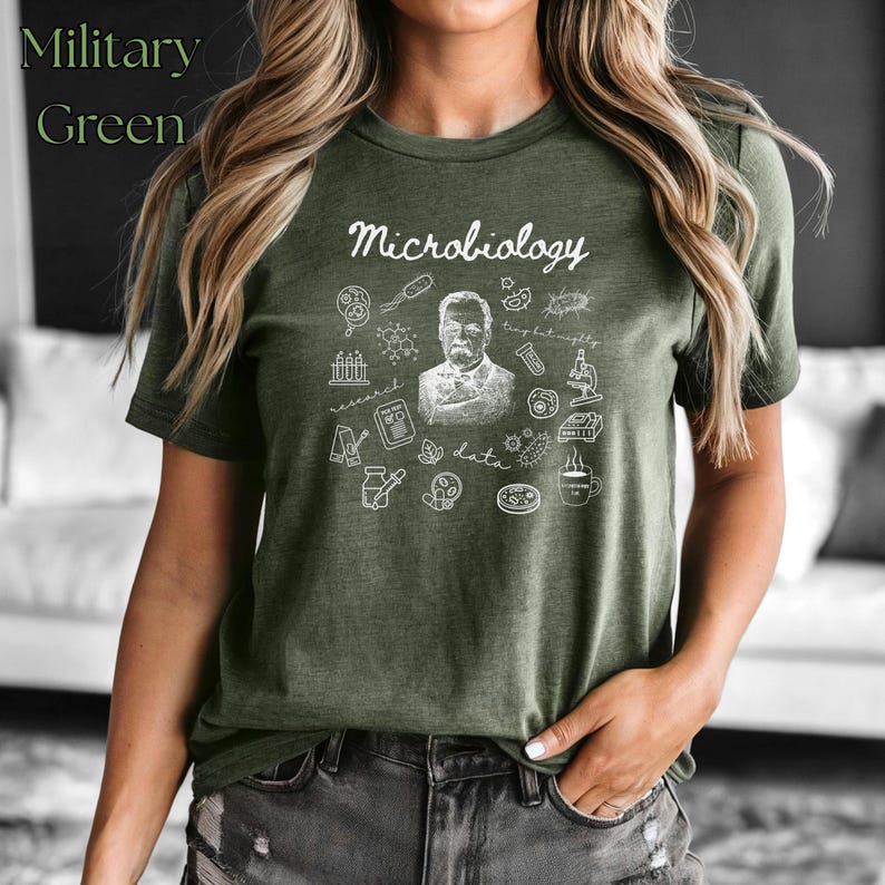May include: Military green t-shirt with the word "Microbiology" in a script font. The shirt features a detailed white graphic of scientific elements, including a portrait, beakers, and a microscope. The shirt is made of a soft material.