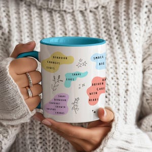 May include: White ceramic mug with a blue interior and handle. The mug features colorful text and illustrations, including phrases like "Behavior Changes Lives" and "Driven with Heart."