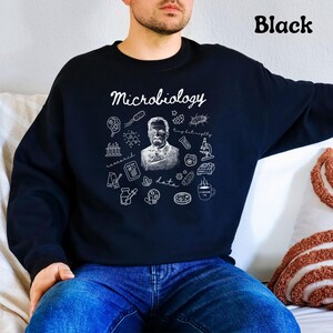 May include: Black sweatshirt with the word "Microbiology" in script. The design includes a portrait, scientific illustrations, and the words "research," "data," and "tiny but mighty" in white. The word "Black" is printed in the upper right corner.