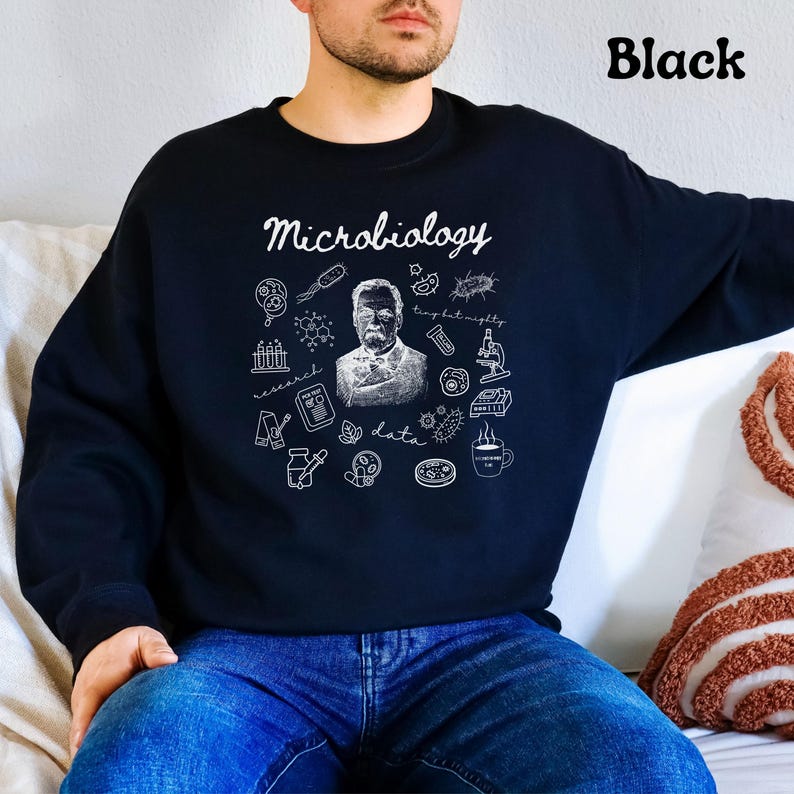 May include: Black sweatshirt with the word "Microbiology" in script. The design includes a portrait, scientific illustrations, and the words "research," "data," and "tiny but mighty" in white. The word "Black" is printed in the upper right corner.
