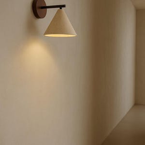 May include: A wall-mounted light fitting with a conical shade. The shade is a light beige colour and is attached to a black arm and a round, dark brown base. The sconce is illuminated, casting a warm glow on the wall.