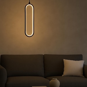 May include: A modern, minimalist pendant light with a black frame and a bright, warm white interior. The light hangs above a dark gray sofa, a wooden coffee table, and a beige pillow. The room has a neutral color palette.