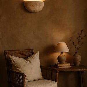 May include: A warm-toned interior scene featuring a wall-mounted sconce, a wooden chair with a linen pillow, and a side table with a lamp and vase. The sconce casts a soft glow, enhancing the room's cozy ambiance.