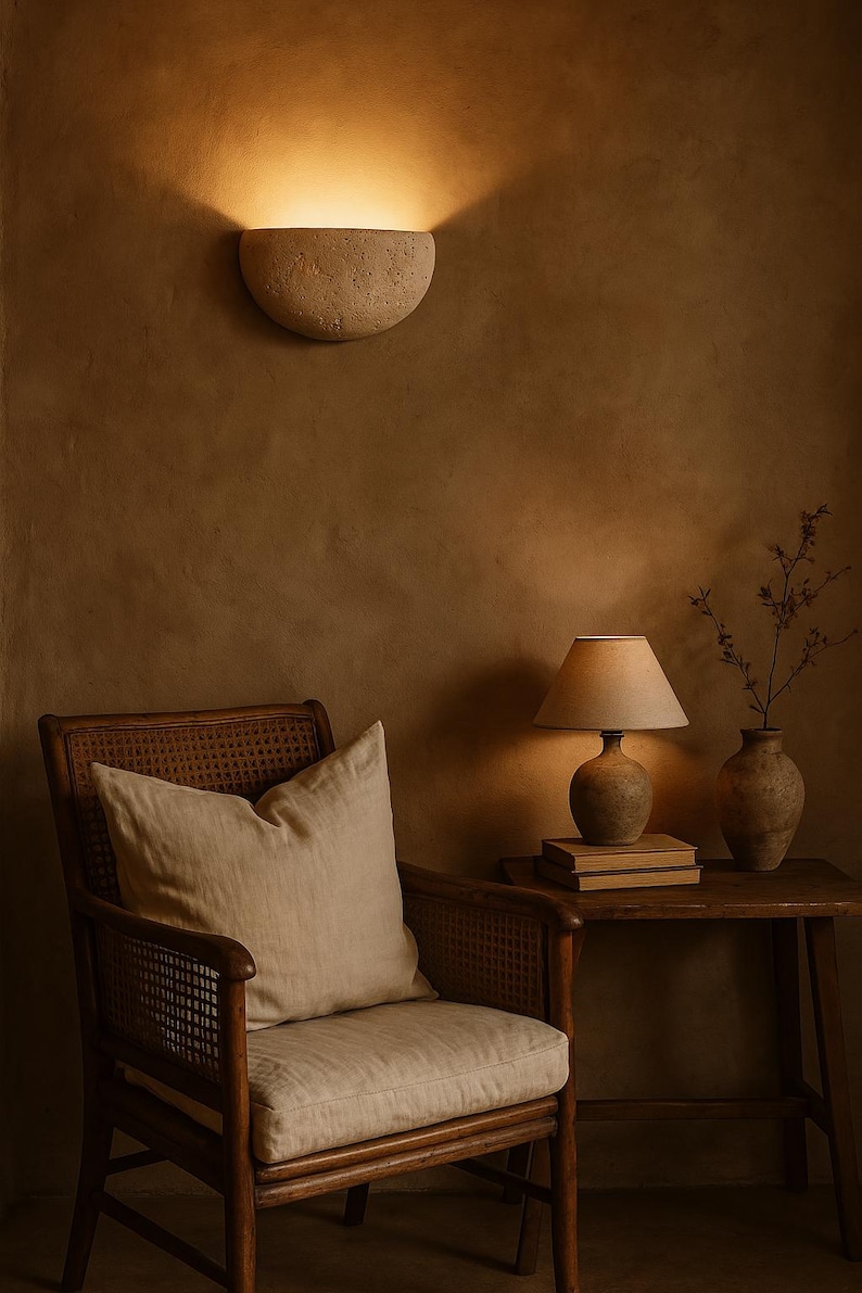 May include: A warm-toned interior scene featuring a wall-mounted sconce, a wooden chair with a linen pillow, and a side table with a lamp and vase. The sconce casts a soft glow, enhancing the room's cozy ambiance.