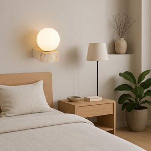 Op de afbeelding: Een moderne slaapkamer met een wandlamp met een bolvormige lamp en een natuurstenen voet. Een nachtkastje met een lamp, een kom en een boek. Een bed met een lichtgekleurd dekbed en kussen.