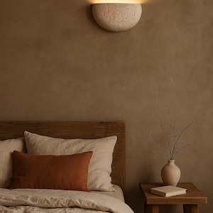 May include: A bedroom scene featuring a warm, neutral color palette. A textured, half-moon shaped wall sconce emits a soft glow. The bed has a wooden headboard, linen bedding, and a rust-colored lumbar pillow. A small wooden side table holds a vase and a book.