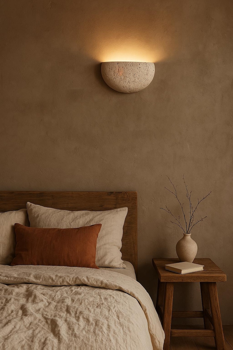 May include: A bedroom scene featuring a warm, neutral color palette. A textured, half-moon shaped wall sconce emits a soft glow. The bed has a wooden headboard, linen bedding, and a rust-colored lumbar pillow. A small wooden side table holds a vase and a book.