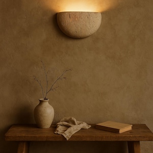 May include: A beige, half-moon shaped wall sconce emits warm light, mounted on a textured brown wall. Below, a wooden bench holds a vase with branches, a folded cloth, and a closed book, creating a serene, minimalist scene.