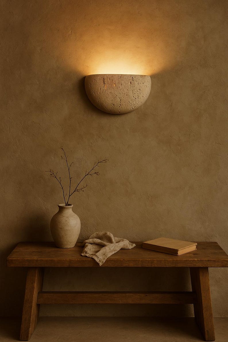 May include: A beige, half-moon shaped wall sconce emits warm light, mounted on a textured brown wall. Below, a wooden bench holds a vase with branches, a folded cloth, and a closed book, creating a serene, minimalist scene.