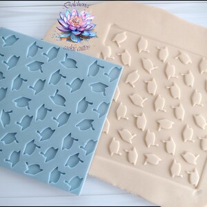 Peut inclure: Un moule en silicone bleu clair avec un motif de chapeaux de graduation, à côté d'une feuille de pâte avec le même motif en relief. L'image montre également une fleur à l'aquarelle et les mots "emporte-pièces".