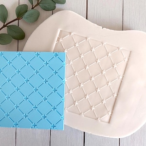 May include: A light blue silicone mold with a crosshatch pattern and a white mold with a similar design. The molds are likely for creating decorative elements, possibly for baking or crafts. The image is set against a light wood background.