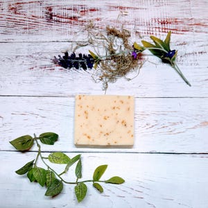 May include: A square bar of soap with a light beige color and small brown flecks throughout. The soap is sitting on a white wooden surface with green leaves and purple flowers on either side.