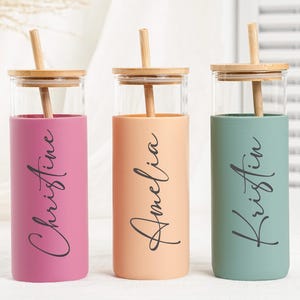 May include: Three personalised glass tumblers with bamboo lids and straws. The tumblers are in shades of pink, peach, and teal, each with a name in elegant script: "Christine", "Amelia", and "Kristin".