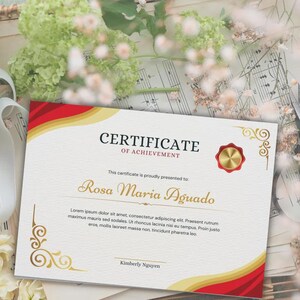 May include: A certificate of achievement with a red and gold border. The certificate reads "Certificate of Achievement" and is presented to Rosa Maria Aguado. The certificate has decorative gold flourishes and a gold seal.
