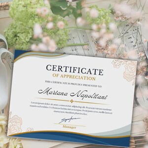 May include: A certificate of appreciation with a blue and gold border. The certificate reads "Certificate of Appreciation" and is presented to "Mariana Napolitani". Decorative floral elements are in the corners. The certificate includes a signature line and the title "Manager".