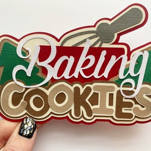 May include: A layered paper cutout with the words "Baking Cookies" in white script and block letters. The design includes a red background, green tree shapes, and a gray whisk, perfect for holiday baking.