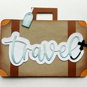 May include: A brown suitcase-shaped paper cutout with the word "Travel" in light blue script. The suitcase has brown straps, orange corner accents, and a light blue luggage tag. A black airplane icon is next to the word "Travel".