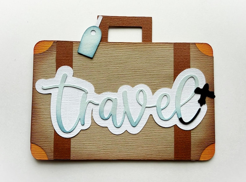 May include: A brown suitcase-shaped paper cutout with the word "Travel" in light blue script. The suitcase has brown straps, orange corner accents, and a light blue luggage tag. A black airplane icon is next to the word "Travel".