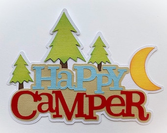 Może przedstawiać: Wycinana papierowa dekoracja z napisem "Happy Camper" niebieskimi i czerwonymi literami na beżowym tle. Wzór zawiera trzy zielone sosny z brązowymi pniami oraz żółty półksiężyc po prawej stronie.