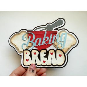 May include: A layered paper craft design with the words "Baking Bread". The design features a red bowl, a whisk, and bread slices. The word "Baking" is in light blue script, and "Bread" is in bold letters with heart accents.