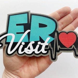 May include: A teal blue and white paper craft with the text "ER Visit" and a red heart with a black outline. The heart is inside a black outline of a heartbeat.