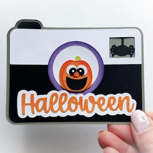 May include: A rectangular paper craft shaped like a camera with a white top and black bottom. It features a central purple circle containing an orange pumpkin with a face, a black spider in the top right corner, and the word Halloween in orange.