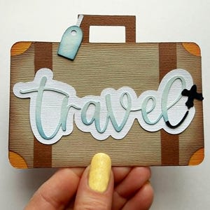 May include: A brown suitcase-shaped paper cutout with the word "Travel" in light blue script. The cutout has brown stripes, orange corner accents, and a blue luggage tag. A black decorative element is attached to the word "Travel".