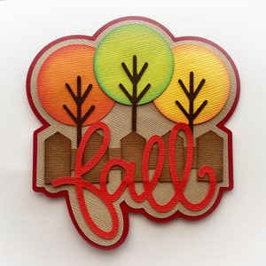Puede incluir: Una tarjeta troquelada con la palabra fall en letras cursivas rojas sobre una valla de madera marr&oacute;n y tres &aacute;rboles con copas circulares texturizadas en naranja, verde lima y amarillo, sobre un fondo beige con borde rojo.