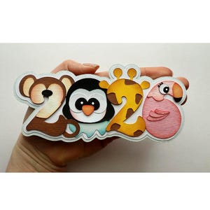 May include: A layered papercraft design spelling out "2026" with animal-themed numbers. The "2" is a monkey, the "0" is a penguin, the second "2" is a giraffe, and the "6" is a flamingo. The design is held in a hand.