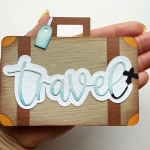 May include: A decorative suitcase-shaped paper cutout with the word "Travel" in a cursive font. The suitcase is tan with brown accents, orange corner details, and a blue luggage tag. A black plus sign is next to the word "Travel".