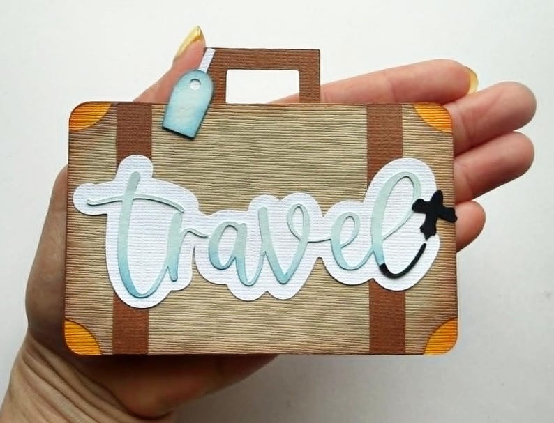 May include: A decorative suitcase-shaped paper cutout with the word "Travel" in a cursive font. The suitcase is tan with brown accents, orange corner details, and a blue luggage tag. A black plus sign is next to the word "Travel".
