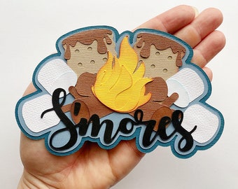 May include: A layered paper craft cutout featuring a bright yellow flame, brown textured logs, tan graham crackers with brown chocolate drips, and white marshmallows. The word S'mores is written in black cursive script across the front.