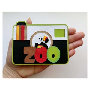 May include: A rectangular paper craft decoration shaped like a camera with a lime green border. The design features a white top section, a black bottom section, a toucan illustration, and the word ZOO in large, textured, multi-colored letters.