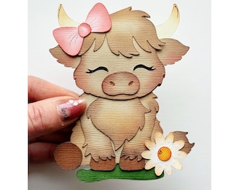 Puede incluir: Artesanía de papel en capas de una vaca de las Tierras Altas con un lazo rosa, ojos cerrados y una margarita. La vaca es marrón con detalles en beige y está sobre una base verde. La artesanía es sostenida por una mano.