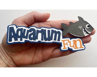 May include: A layered paper cutout with the words "Aquarium Fun" in blue and orange. A gray shark with white teeth and a smiling face is next to the text. The cutout is held in a hand.