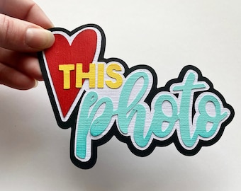 May include: A teal blue and white paper cutout with the words "THIS photo" in a bold, cursive font. A red heart with a yellow "THIS" inside is on the left side of the cutout.