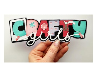 May include: A colorful die-cut design with the words "Crafty Girls". The letters are in various colors, including teal, pink, and red, with craft-themed embellishments like scissors and a button. The word "girls" is in a cursive font.