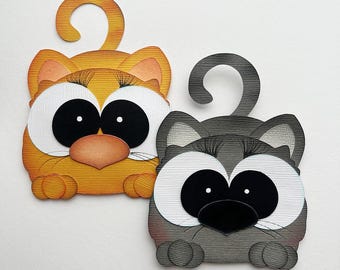 May include: Two whimsical cat-shaped paper cutouts. One is orange with peach accents, large white eyes, and a black nose. The other is gray with black accents, similar eyes, and a black nose. Both have curled tails and are designed with textured paper.
