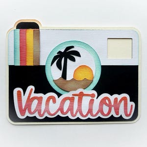 May include: A decorative camera-shaped item with a black body and a white top. The design includes a palm tree and sunset graphic inside a circle, and the word "Vacation" in a script font. The camera has a colorful striped detail on the side.