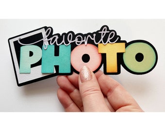 May include: A die-cut paper embellishment featuring the word "PHOTO" in large, colorful block letters with a gradient finish, topped with the word "favorite" in a white script font, all set against a black and white square frame.