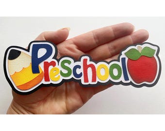 May include: A colorful paper craft with the word "Preschool" in large letters. The letters are decorated with different colors and textures. A red apple with a green leaf and a yellow and orange pencil are on either side of the word.